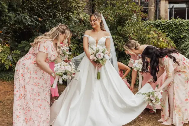 A bride in a white gown holds a bouquet while four bridesmaids in pink floral dresses help arrange her dress outdoors in the gardens of Whirlow Brook Hall.