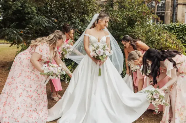 A bride in a white gown and veil stands outdoors at Whirlow Brook Hall, holding a bouquet, while five bridesmaids in pink floral dresses adjust her dress and veil, surrounded by lush greenery.