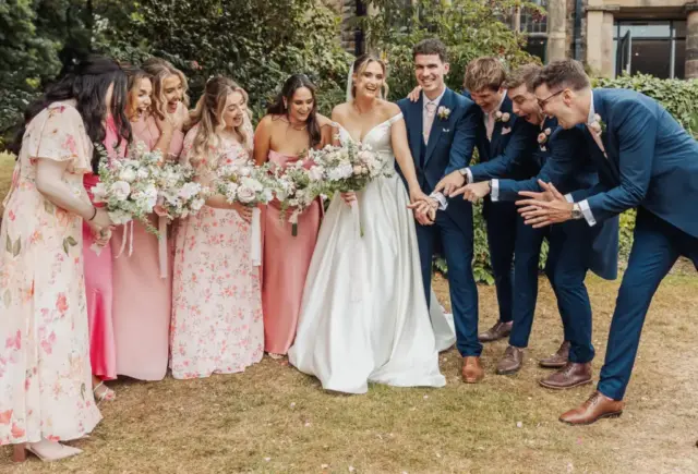 A bride in a white dress stands smiling outdoors at Whirlow Brook Hall, surrounded by bridesmaids in pink dresses and groomsmen in blue suits, all laughing and pointing toward the bride amid lush greenery.
