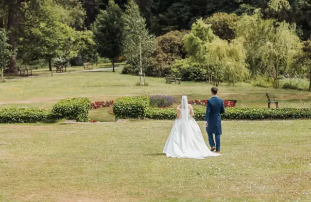 A bride in a white dress and a groom in a blue suit walk together across the green lawn at Whirlow Brook Hall, surrounded by trees, bushes, and flowers on their wedding day.