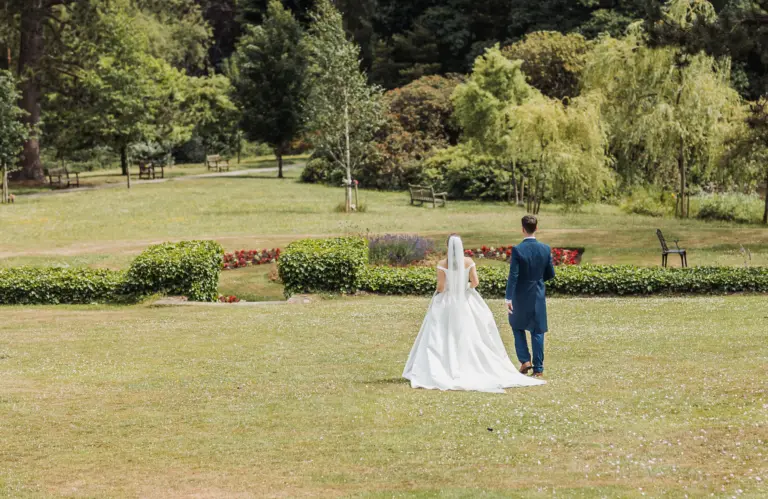 A bride in a white dress and a groom in a blue suit walk together across the green lawn at Whirlow Brook Hall, surrounded by trees, bushes, and flowers on their wedding day.