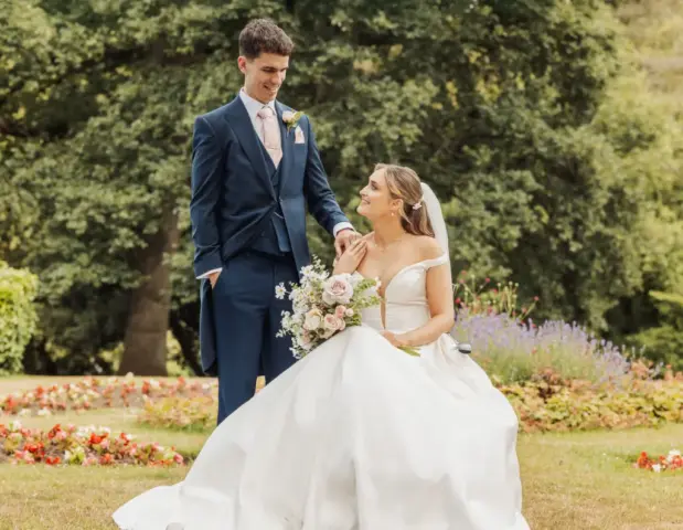 A bride in a white gown holds a bouquet as she sits on the grass at Whirlow Brook Hall, smiling up at her groom in a navy suit, surrounded by colorful flowers and trees in the background.
