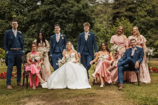 A wedding party poses outdoors on grass at Whirlow Brook Hall, with the bride in a white gown and groom in blue at the center, surrounded by smiling bridesmaids in pink dresses and groomsmen in blue suits. Trees and flowers form a lovely backdrop.