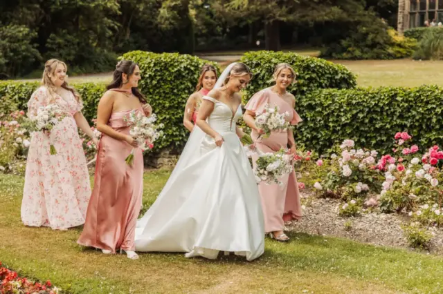 A bride in a white gown walks outdoors with four bridesmaids in pink dresses, holding bouquets, surrounded by blooming flowers and greenery at Whirlow Brook Hall.