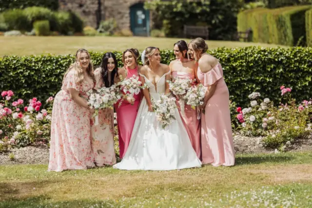 A bride in a white gown stands with five bridesmaids in pink dresses, all smiling and holding bouquets, amid blooming flowers and greenery at Whirlow Brook Hall.