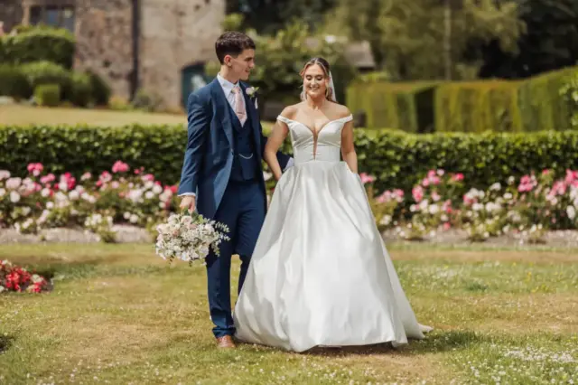 A bride in a white gown and a groom in a blue suit walk together in the gardens of Whirlow Brook Hall, surrounded by blooming flowers and greenery, smiling at each other on their wedding day. The groom holds a bouquet of flowers.