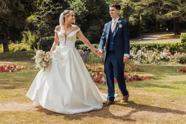 A bride in a white gown and a groom in a navy suit hold hands and smile at each other while walking on a sunny lawn surrounded by colorful flowers and greenery at Whirlow Brook Hall.