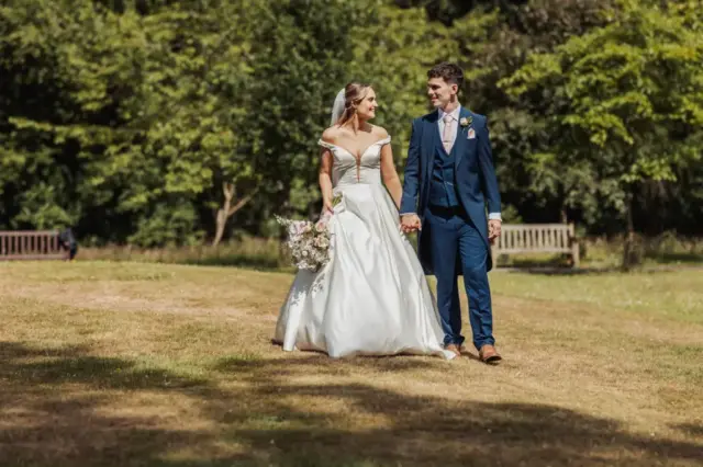 A bride in a white dress holding a bouquet and a groom in a blue suit walk hand in hand on grass at Whirlow Brook Hall’s sunny park, smiling at each other. Trees and benches are visible in the background.