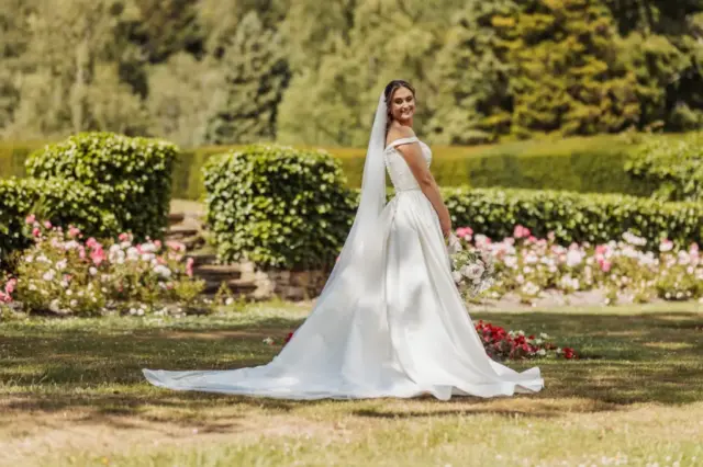 A bride in a flowing white gown and long veil stands outdoors at Whirlow Brook Hall, holding a bouquet and smiling over her shoulder, surrounded by green hedges and colorful flowers on her wedding day.