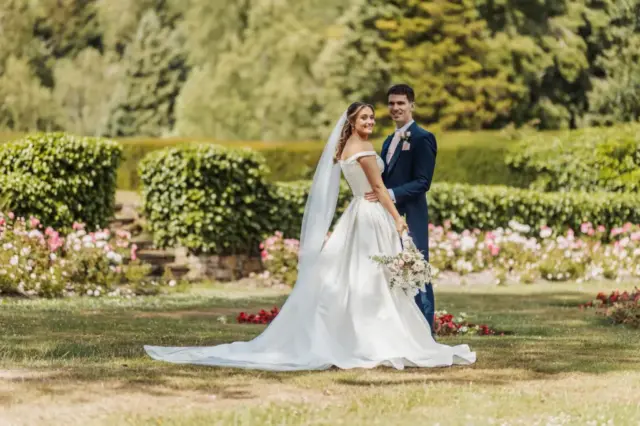 A bride in a white wedding dress and veil stands with her groom in a navy suit at Whirlow Brook Hall, both smiling and holding a bouquet, surrounded by garden greenery and blooming flowers.