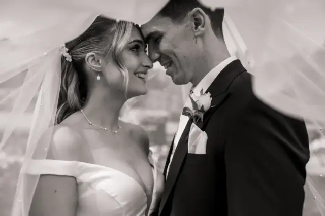 A bride and groom stand close together under a veil, smiling lovingly at each other on their wedding day at Whirlow Brook Hall. The black and white image captures an intimate and joyful moment between the wedding couple.