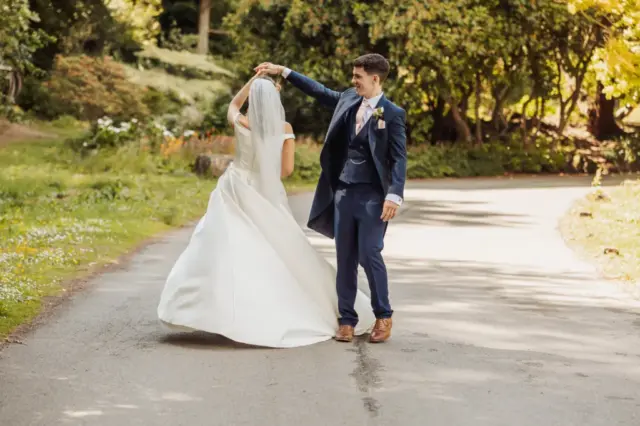 A groom in a navy suit twirls his bride, dressed in a white gown and veil, on a tree-lined path at Whirlow Brook Hall in a sunlit outdoor wedding setting.