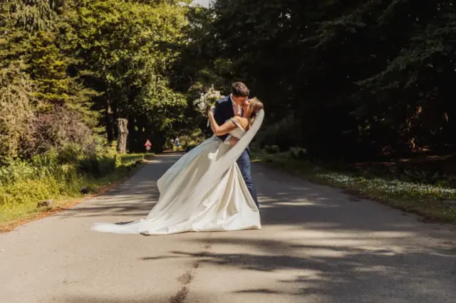 A bride and groom share a romantic kiss in the middle of a sunlit road near Whirlow Brook Hall, surrounded by trees. Her long white wedding dress and veil flow behind her as the groom gently dips her backward.