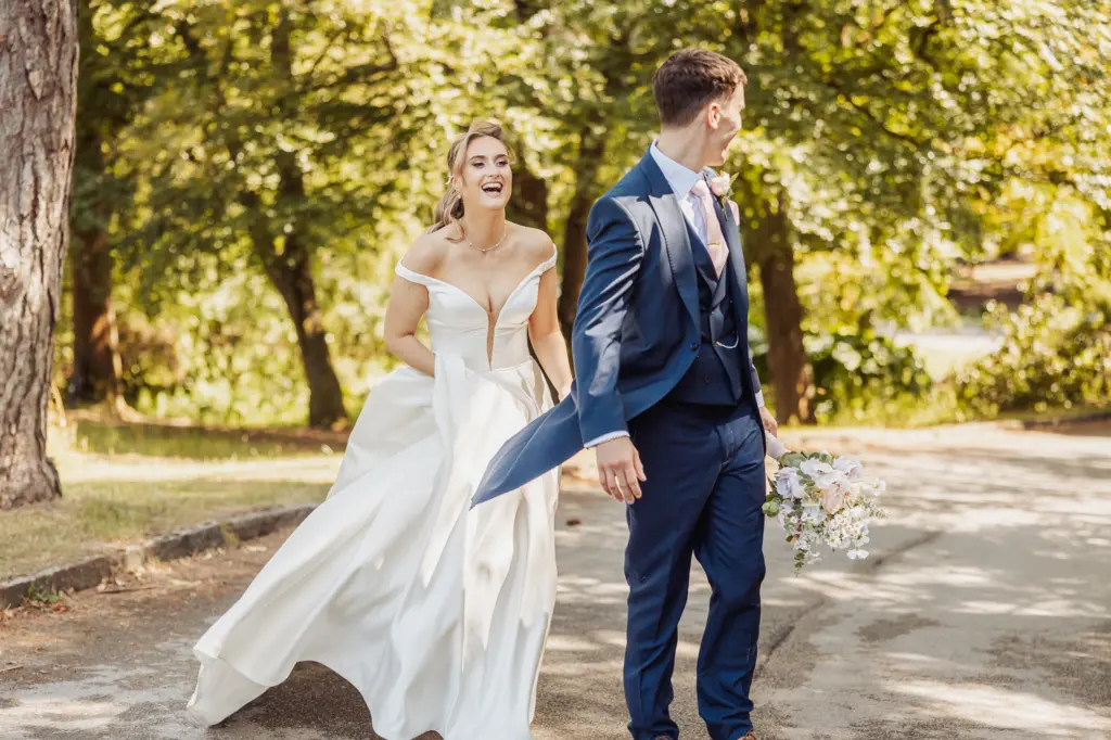 A bride in a white dress smiles and walks hand in hand with her groom, holding a bouquet, outdoors on a sunny day with trees in the background at Whirlow Brook Hall.