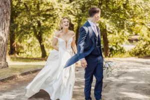 A bride in a white dress smiles and walks hand in hand with her groom, holding a bouquet, outdoors on a sunny day with trees in the background at Whirlow Brook Hall.