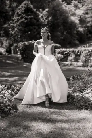 A bride in an off-the-shoulder, flowing white gown walks through a sunny garden at Whirlow Brook Hall, holding up her dress. Trees and bushes surround her as she looks ahead, sunlight casting soft shadows.