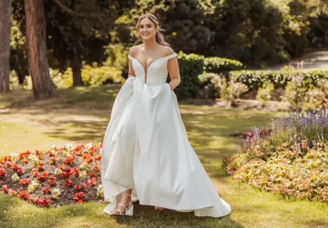 A smiling bride in an off-the-shoulder white wedding gown holds her dress while walking through a sunny garden at Whirlow Brook Hall, with colorful flowers and greenery in the background.