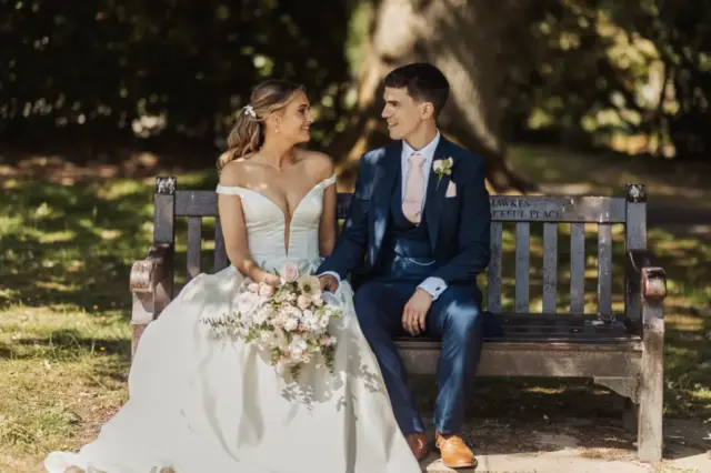 A bride in a white dress and groom in a blue suit sit together on a wooden bench outdoors at Whirlow Brook Hall, holding hands and looking at each other, with a bouquet of flowers in the bride’s lap—a perfect wedding moment.