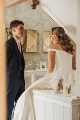 A bride in an off-shoulder white gown sits on a counter, facing her groom in a navy suit. They gaze at each other in a softly lit, elegant room adorned with framed photos and decorative mirrors at Whirlow Brook Hall.