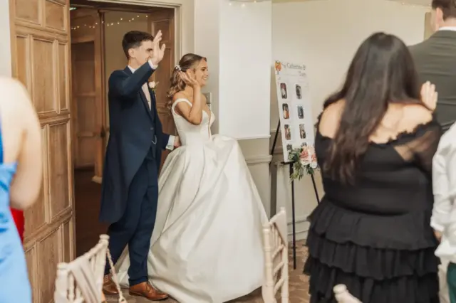 A bride and groom enter their Whirlow Brook Hall wedding reception, smiling and waving at guests. The bride wears a white gown, and the groom is in a dark suit. Guests stand to greet them; a seating chart is visible in the background.