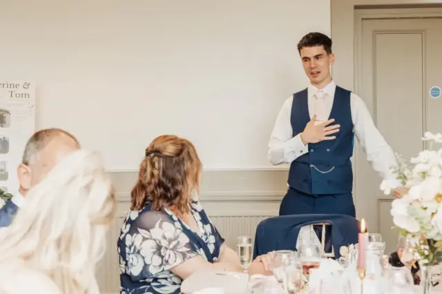 A young man in a blue vest and tie stands, giving a speech with one hand on his chest, while seated guests at Whirlow Brook Hall listen attentively at a formal wedding reception, possibly addressing the bride and groom.