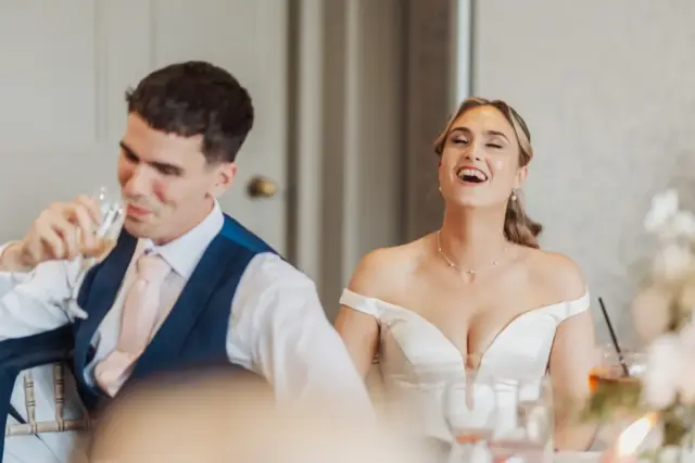 A smiling bride in an off-shoulder white dress laughs joyfully next to a man in a blue suit and pale pink tie, who is sipping from a glass at their wedding reception at Whirlow Brook Hall.