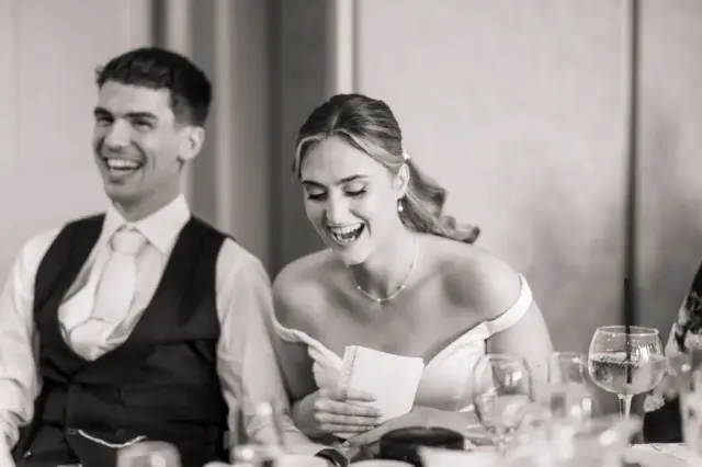 A bride and groom sit side by side at a table, both laughing joyfully at their Whirlow Brook Hall wedding. The bride, in an off-shoulder dress, holds a notepad while the groom in a suit and tie smiles beside her. Glasses and tableware are visible in the foreground.