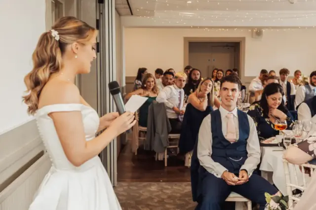 At a Whirlow Brook Hall wedding reception, the bride in a white dress gives a heartfelt speech with a microphone and notes, while the groom, dressed in a suit, listens attentively among the seated guests.