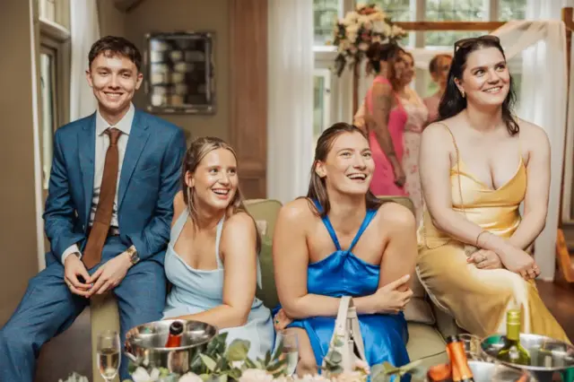 Four young adults in formal attire sit smiling on a couch at a bright Whirlow Brook Hall wedding. Three women in pastel dresses and one man in a blue suit are in the foreground, with more guests and floral decor in the background.