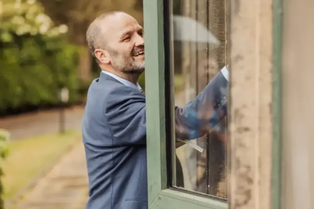A man in a blue suit smiles as he reaches through an open window from the outside at Whirlow Brook Hall, with lush greenery in the background—perfect for a charming wedding moment.