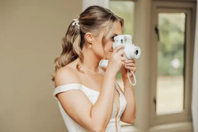 A bride in an off-the-shoulder white dress holds a white instant camera up to her face, capturing a candid moment indoors near large windows at Whirlow Brook Hall.