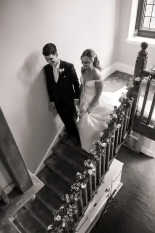 A bride and groom walk down the flower-decorated staircase at Whirlow Brook Hall, holding hands. The bride wears an off-shoulder white gown as sunlight streams through a window, creating a warm, intimate wedding atmosphere.