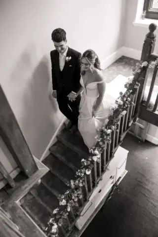 A bride and groom walk down a decorated staircase at Whirlow Brook Hall, holding hands and smiling. The bride wears an off-the-shoulder gown; the groom is in a suit with a bow tie. The timeless scene feels like a classic wedding captured in black and white.
