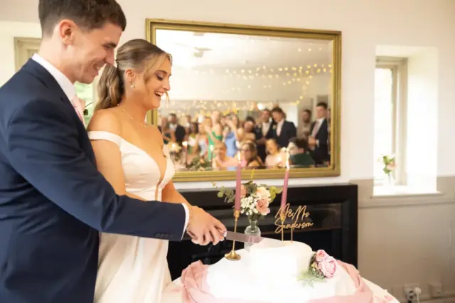 A bride and groom smile as they cut their wedding cake together at Whirlow Brook Hall. The cake is decorated with pink flowers and a Mr & Mrs topper, while guests are reflected in a large mirror behind them, watching and celebrating.