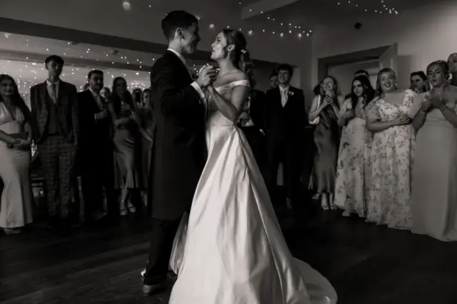 A bride and groom share their first dance in a softly lit room at Whirlow Brook Hall, surrounded by smiling guests in formal attire; string lights hang overhead, creating a warm, celebratory wedding atmosphere.