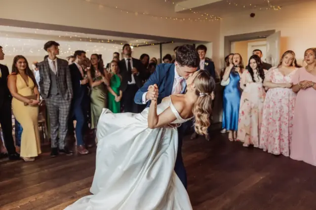 A bride and groom share a romantic dance at Whirlow Brook Hall while the groom dips the bride. Surrounded by elegantly dressed guests in a warmly lit room adorned with string lights, they celebrate their wedding with joy.