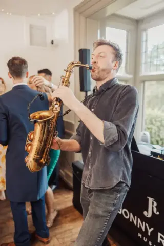 A man plays a saxophone indoors near a DJ booth at Whirlow Brook Hall, with wedding guests dancing in the background. He is wearing a casual shirt and jeans as sunlight streams through large windows behind him.