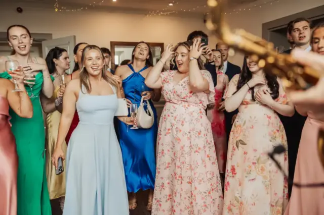 A group of people dressed in colorful formal attire stand together indoors at Whirlow Brook Hall, many smiling and reacting with excitement as a saxophonist performs. The scene captures the lively atmosphere of a celebratory wedding event.