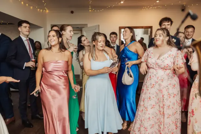 A group of people dressed in colorful formal attire, including suits and dresses, stand together indoors at a lively wedding at Whirlow Brook Hall, smiling and chatting. Some hold drinks, and fairy lights decorate the ceiling.
