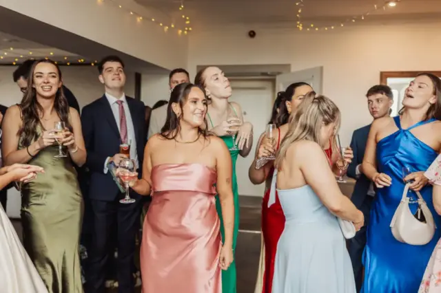 A group of well-dressed people stand indoors at a wedding celebration at Whirlow Brook Hall, holding drinks and smiling. Women wear colorful dresses, men wear suits, and string lights hang from the ceiling, creating a festive atmosphere.
