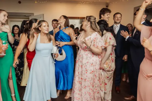 A group of people in formal and colorful dresses and suits are standing closely together indoors at Whirlow Brook Hall, smiling and talking at what appears to be a wedding reception. Some are holding drinks, celebrating with the bride.