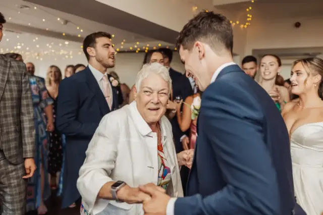 An elderly woman and a young man, both smiling, dance together at a lively wedding at Whirlow Brook Hall, with guests dressed formally and warm string lights overhead.