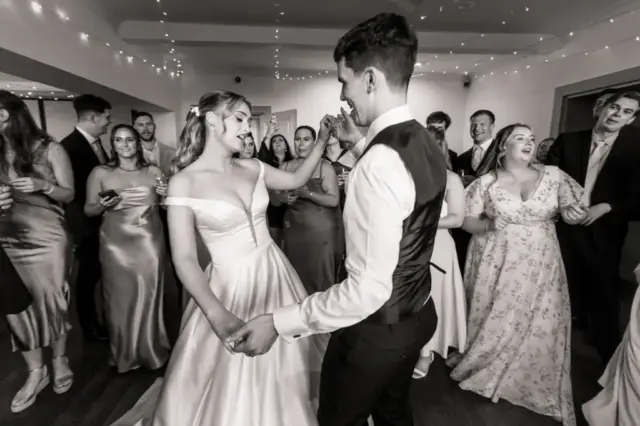 A bride and groom dance together, smiling and holding hands, surrounded by guests in formal attire who are watching and celebrating at their Whirlow Brook Hall wedding. The scene is lively and joyful, with string lights above.