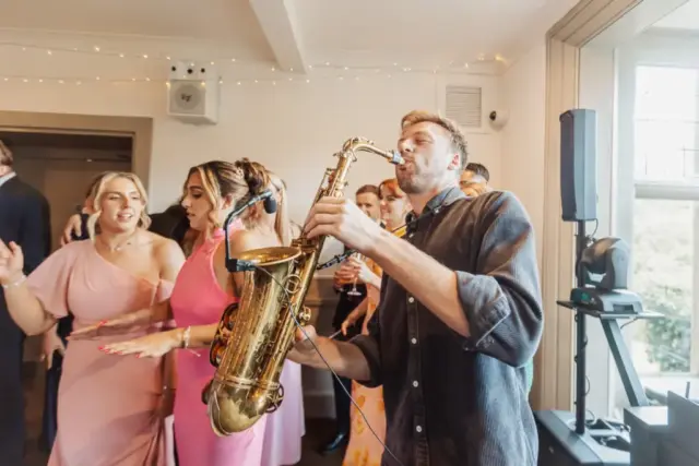 A man plays a saxophone at an indoor wedding party at Whirlow Brook Hall, surrounded by guests dancing in elegant attire, with women in pink dresses and a bright, festive atmosphere celebrating the bride.