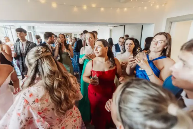 A group of people in formal attire are gathered closely together at a wedding reception at Whirlow Brook Hall, dancing and socializing under string lights. Some hold drinks and appear to be enjoying themselves.