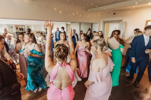 A group of people dressed in formal attire dance and celebrate at a Whirlow Brook Hall wedding, with women in colorful dresses and men in suits, under string lights and surrounded by tables as the bride joins the festivities.