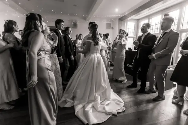 A bride in a long, elegant gown stands surrounded by guests in formal attire at Whirlow Brook Hall, all smiling and chatting in a bright, decorated room during the joyful wedding celebration.