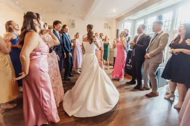 A bride in a white dress dances joyfully at the center of Whirlow Brook Hall, surrounded by smiling guests in formal attire, all clapping and celebrating at this bright indoor wedding reception.
