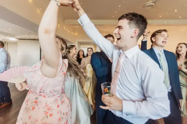 A group of people in formal attire dance and celebrate indoors, smiling and laughing. At this Whirlow Brook Hall wedding, a man in a white shirt and tie twirls the bride in a pink floral dress holding a fan, as others enjoy the lively atmosphere.
