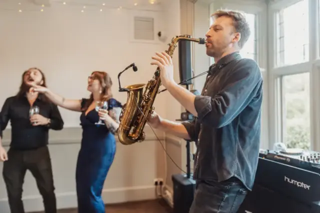 At Whirlow Brook Hall, a man plays the saxophone indoors while a bride in a blue dress and another man sing and dance behind him, holding drinks. Warm lighting and a DJ setup add to the lively wedding atmosphere in the background.